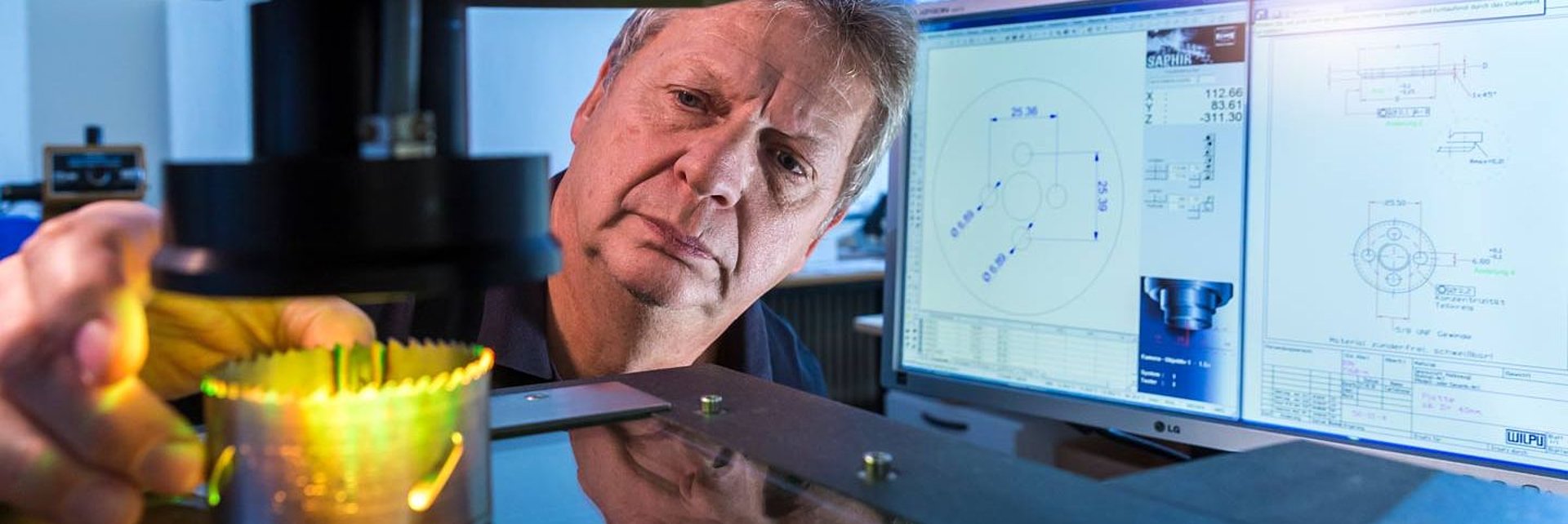 Mann fokussiert auf Lochsäge neben Computerbildschirmen mit technischen Zeichnungen. Mann fokussiert auf Lochsäge neben Computerbildschirmen mit technischen Zeichnungen.